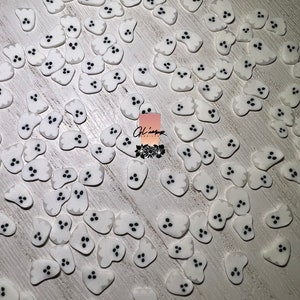 May include: A large quantity of white ghost-shaped polymer clay charms with black eyes. The charms are scattered on a white wooden surface.