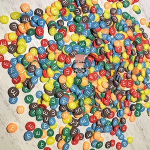 May include: A large pile of colorful round candy sprinkles with the letter "M" printed in white on each sprinkle. The sprinkles are in shades of red, orange, yellow, green, blue, and brown.
