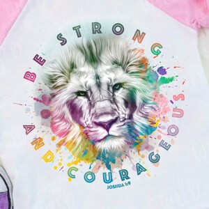 Pode incluir: Uma camiseta de beisebol branca e rosa com um gráfico de leão colorido e o texto "Seja forte e corajoso Josué 1:9".