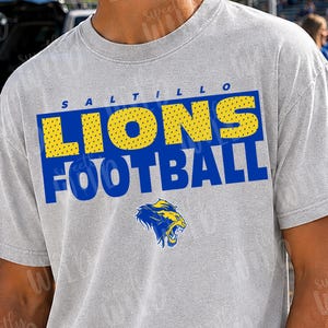 May include: A light gray short-sleeve t-shirt featuring the text Saltillo Lions Football in blue and yellow. The word Lions is in yellow with a blue outline, and the word Football is in blue. A blue and yellow lion head logo is centered below.