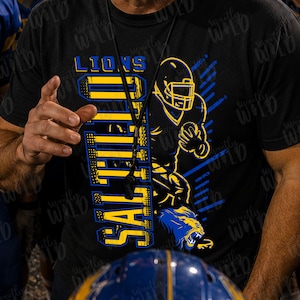 May include: A black short-sleeved t-shirt featuring a graphic print with the text LIONS and SALFIELD in yellow and blue. The design includes a stylized illustration of a football player in a helmet and a roaring lion head in blue and yellow.