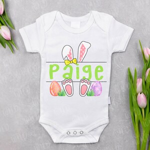 May include: White baby onesie featuring a green Easter bunny design. The bunny has pink ears and paws, a yellow bow tie, and is holding a sign that says "Paige". There are three colourful Easter eggs on the ground.