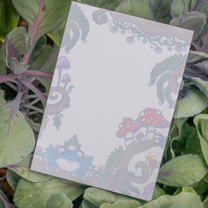 May include: A white notepad with a whimsical design featuring mushrooms, ferns, and a frog in shades of red, blue, purple, and green. The notepad is surrounded by green and purple cabbage leaves.