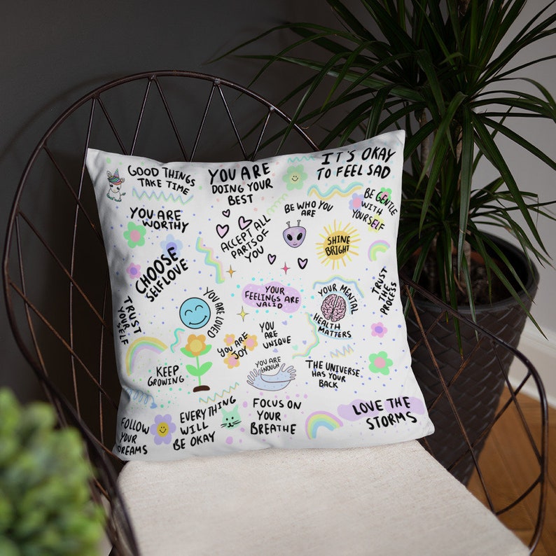 K&ouml;nnte beinhalten: Ein wei&szlig;es Kissen mit bunten Illustrationen und positiven Affirmationen, darunter "Du bist es wert", "W&auml;hle Selbstliebe", "Wachse weiter", "Liebe die St&uuml;rme" und "Es ist okay, traurig zu sein".
