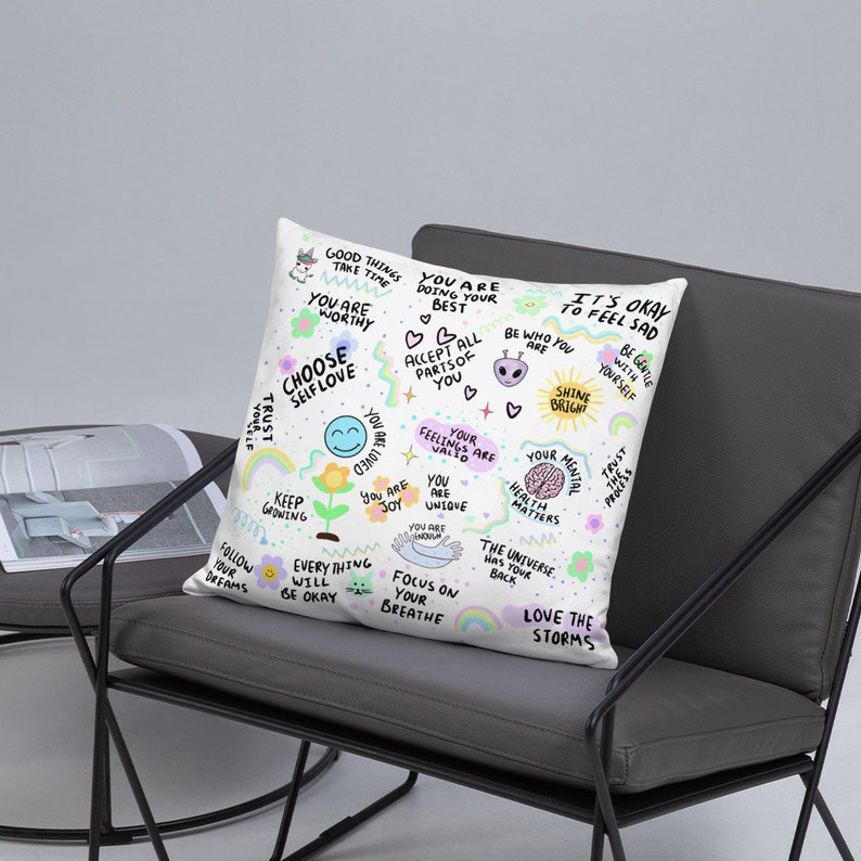 K&ouml;nnte beinhalten: Ein wei&szlig;es Kissen mit buntem Text und Illustrationen. Der Text enth&auml;lt S&auml;tze wie "W&auml;hle Selbstliebe", "Du bist es wert", "Es ist okay, sich traurig zu f&uuml;hlen", "Strahle", "Liebe die St&uuml;rme" und "Das Universum hat deinen R&uuml;cken".