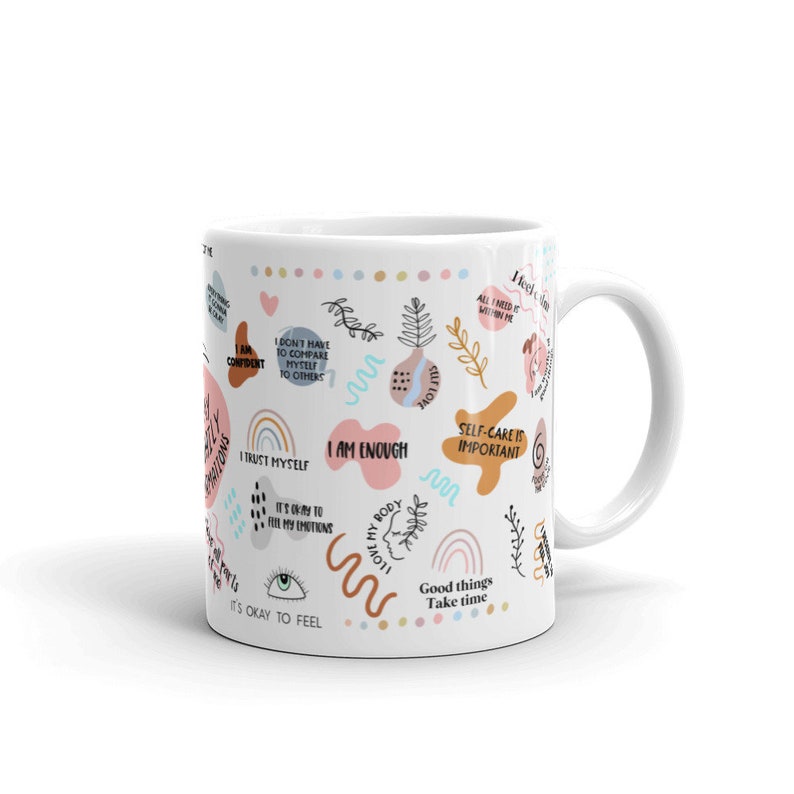 Puede incluir: Taza de cer&aacute;mica blanca decorada con frases y garabatos inspiradores de colores. La taza presenta frases como "Soy suficiente", "El autocuidado es importante" y "Est&aacute; bien sentir". El dise&ntilde;o incluye arco&iacute;ris, corazones y elementos florales.