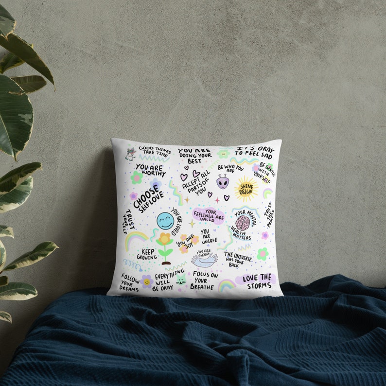 K&ouml;nnte beinhalten: Ein wei&szlig;es Kissen mit einem farbenfrohen Design mit positiven Affirmationen und Illustrationen. Der Text enth&auml;lt S&auml;tze wie "Du bist es wert", "W&auml;hle Selbstliebe", "Wachse weiter", "Liebe die St&uuml;rme" und "Strahle hell".