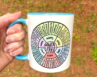 May include: White ceramic mug with a blue rim and handle. The mug features a colorful wheel with the words "How Do I Feel?" in the center and a variety of feelings listed around the wheel.