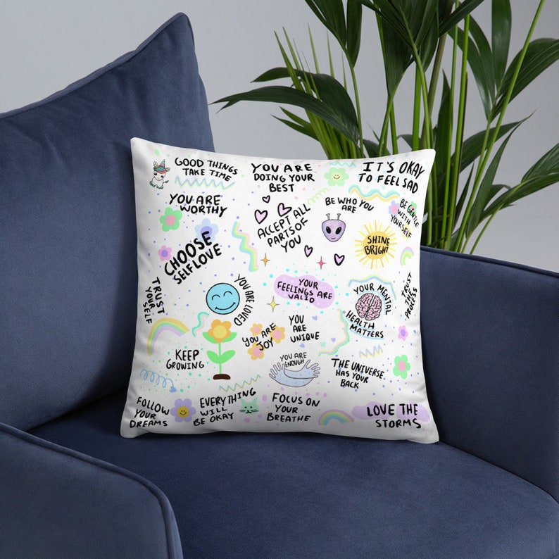K&ouml;nnte beinhalten: Ein wei&szlig;es Kissen mit bunten Illustrationen und positiven Affirmationen. Das Kissen zeigt S&auml;tze wie "W&auml;hle Selbstliebe", "Du bist es wert", "Wachse weiter" und "Liebe die St&uuml;rme".