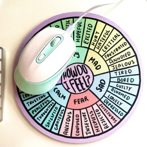 May include: A round mouse pad with a colorful wheel featuring the words "How Do I Feel?" and various emotions. The wheel is divided into sections with different colors and emotions written in black. A white computer mouse with a light blue bottom is on top of the mouse pad.