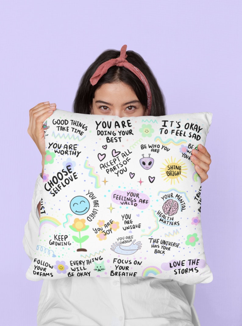 K&ouml;nnte beinhalten: Wei&szlig;es Kissen mit bunten handgezeichneten Illustrationen und positiven Affirmationen, darunter "W&auml;hle Selbstliebe", "Du bist es wert", "Es ist okay, sich traurig zu f&uuml;hlen", "Strahle hell", "Liebe die St&uuml;rme" und "Das Universum hat deinen R&uuml;cken".