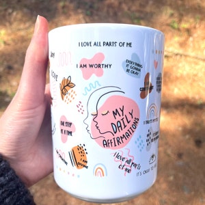 Puede incluir: Taza de cer&aacute;mica blanca con el texto "My Daily Affirmations" y otras afirmaciones positivas en varios colores. La taza presenta un dise&ntilde;o de un rostro femenino y formas abstractas coloridas.