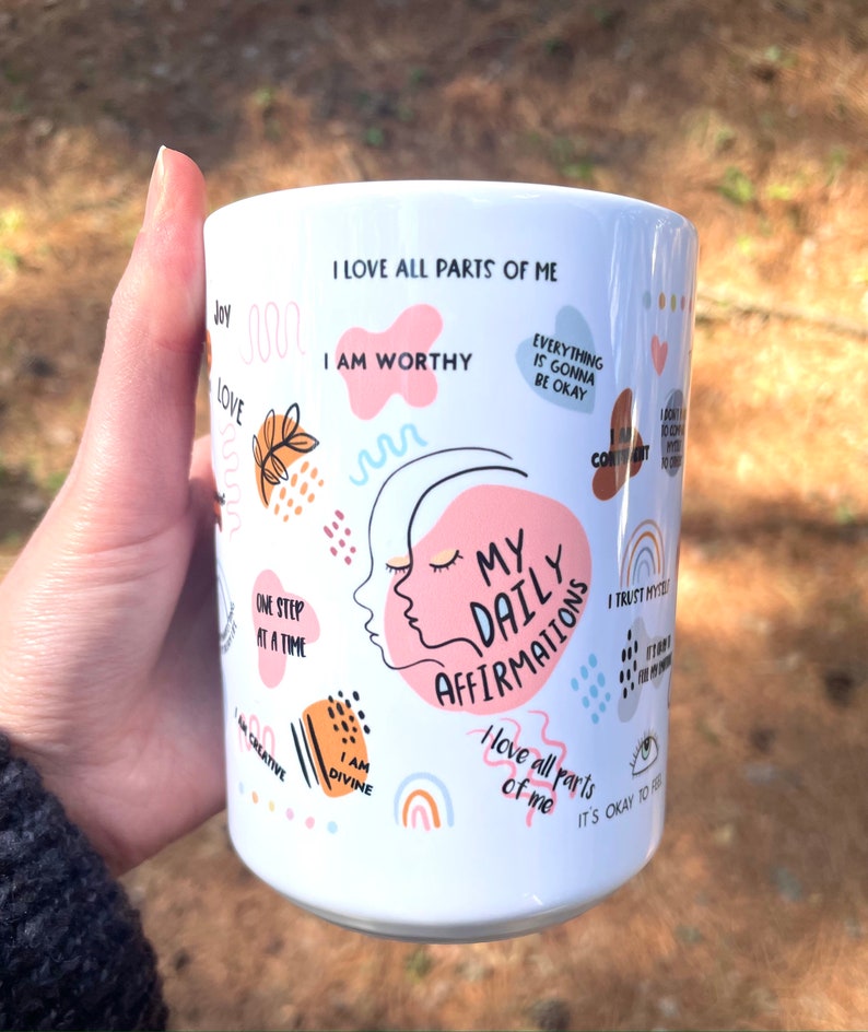 Puede incluir: Taza de cer&aacute;mica blanca con el texto "My Daily Affirmations" y otras afirmaciones positivas en varios colores. La taza presenta un dise&ntilde;o de un rostro femenino y formas abstractas coloridas.