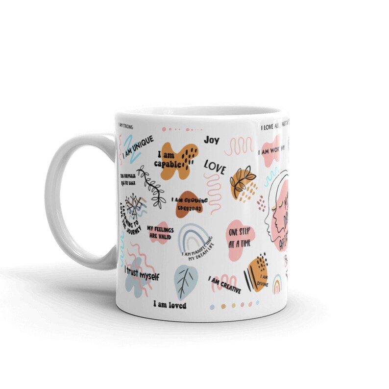 Puede incluir: Taza de cer&aacute;mica blanca con asa curva, decorada con afirmaciones coloridas y formas abstractas. La taza presenta frases como &laquo;Soy &uacute;nico&raquo;, &laquo;Soy capaz&raquo; y &laquo;Soy amado&raquo;, junto con otras afirmaciones positivas.