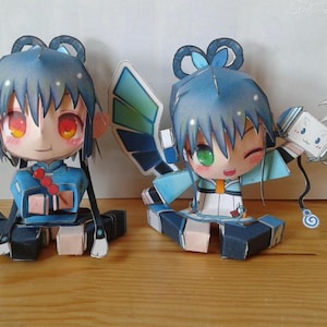 May include: Two papercraft anime-style figures. One figure is seated, wearing a blue outfit with red accents and holding a black staff. The other figure is winking, wearing a blue and white outfit, and holding a papercraft object.