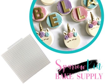 Puede incluir: Un conjunto de galletas decoradas con temática de unicornio. Las galletas están decoradas con glaseado rosa, morado y azul y letras doradas que deletrean "BELLA". También hay una estera de hornear de silicona blanca cuadrada con líneas en relieve.