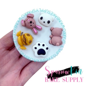 May include: A white silicone mould with six different animal shapes: a frog, a paw print, a bunny, a bear, a cat, and a bird. The mould is on a white surface.