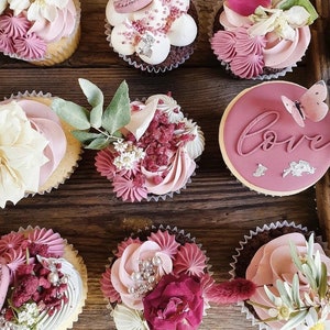 May include: Assortment of cupcakes decorated with pink frosting, flowers, and sprinkles. Some cupcakes have the word "love" written in pink frosting on top.