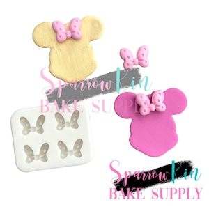 Peut inclure: Un moule en silicone avec quatre cavités en forme de nœuds à pois. Il y a deux formes de tête de souris rose et or avec des nœuds sur le dessus. Le texte "Sparrow Kin BAKE SUPPLY" est visible sur l'image.