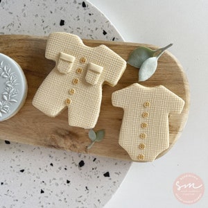 May include: Two sugar cookies decorated with icing to look like baby clothes. One cookie is shaped like a onesie and the other is shaped like a short-sleeved shirt. Both cookies have a checkered pattern and gold buttons.