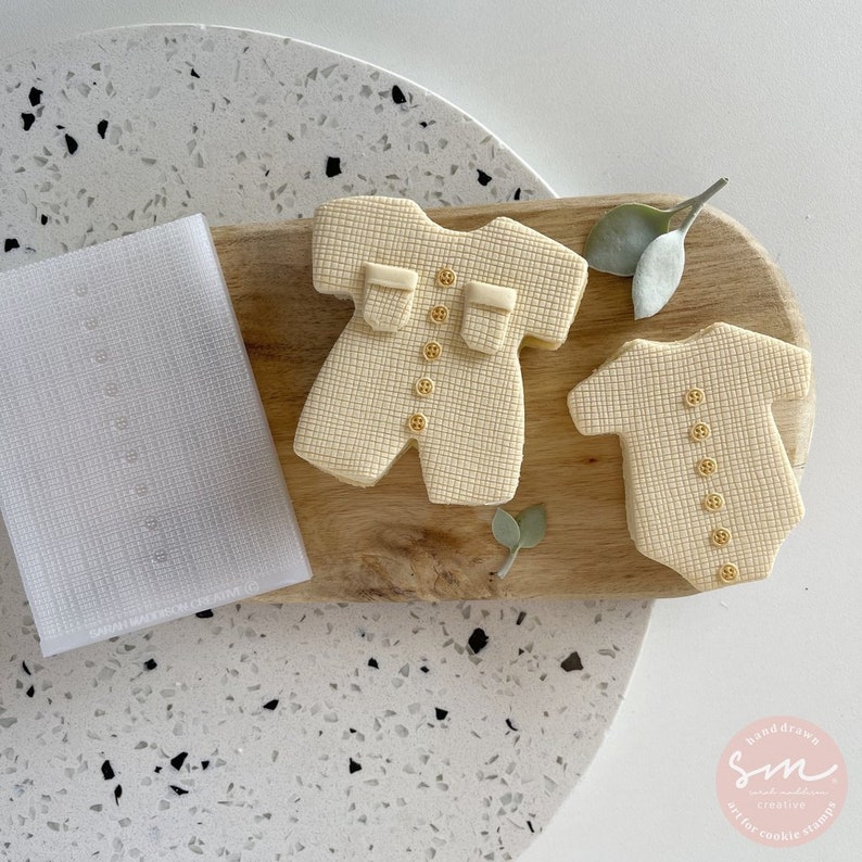 May include: Two beige cookie cutters shaped like baby onesies, one with long sleeves and one with short sleeves. The cutters are on a wooden cutting board with green leaves. The cutters have a textured surface and gold buttons.