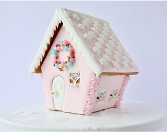 May include: A pink gingerbread house decorated with white icing, candy, and a wreath. The roof is covered in white icing with a snowflake design. The house has a white door with a gold knob and two windows with white lace curtains.