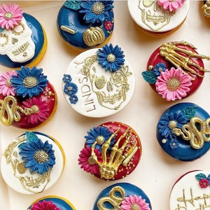 Pode incluir: Uma variedade de donuts decorados com o tema Dia dos Mortos. Os donuts apresentam desenhos de caveiras, cobras e m&atilde;os de esqueleto, juntamente com detalhes florais azuis e rosa. Alguns t&ecirc;m o nome "Lindsay". Cores douradas e brancas proeminentes.