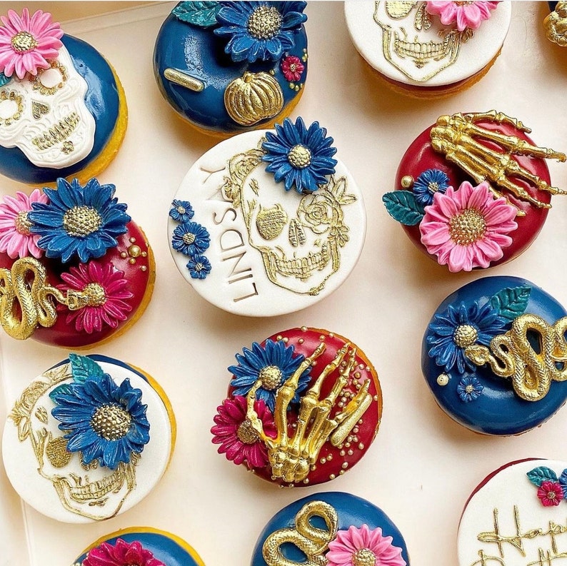 Pode incluir: Uma variedade de donuts decorados com o tema Dia dos Mortos. Os donuts apresentam desenhos de caveiras, cobras e m&atilde;os de esqueleto, juntamente com detalhes florais azuis e rosa. Alguns t&ecirc;m o nome "Lindsay". Cores douradas e brancas proeminentes.