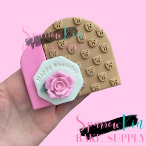 May include: A brown silicone cake decorating stamp with a repeating pattern of bear faces. The stamp is attached to a pink and white octagonal cake topper with the text "Happy Birthday" and a pink rose.
