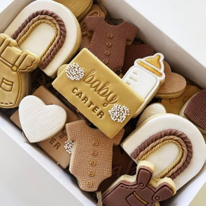 May include: A box of decorated sugar cookies with various baby-themed designs, including a onesie, a bottle, a heart, and a sign that reads "baby carter".