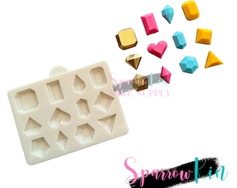 Puede incluir: Un molde de silicona blanco con 12 formas diferentes de gemas, incluyendo corazones, diamantes y cuadrados. El molde se utiliza para hacer decoraciones de fondant o pasta de goma para pasteles y cupcakes. También hay varias golosinas de colores en forma de gemas esparcidas alrededor del molde.