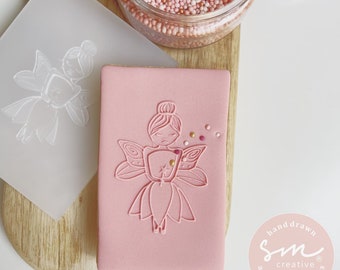 Op de afbeelding: Een roze koekje met een witte omtrek van een fee met vleugels en een toverstaf. Het koekje is versierd met roze en witte sprinkles. De afbeelding toont ook een doorzichtige plastic koekjesstempel met een fee-ontwerp.