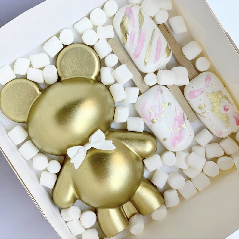 Pode incluir: Um doce dourado em forma de urso com um la&ccedil;o branco, rodeado por marshmallows e tr&ecirc;s picol&eacute;s decorados em branco e rosa com detalhes dourados, tudo dentro de uma caixa branca.