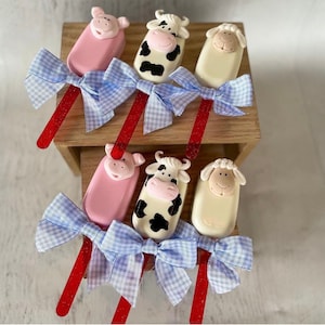 May include: Six farm animal-themed popsicles. The popsicles are shaped like a pig, cow, and sheep. Each popsicle has a red glitter stick and a blue gingham bow. The popsicles are arranged on a wooden display.
