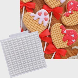 Può includere: Uno stampo in silicone quadrato bianco con una griglia di piccole rientranze piramidali, posizionato accanto a diversi biscotti a forma di cuore su bastoncini. I biscotti sono decorati con glassa rossa o rosa chiaro e piccoli cuori bianchi.