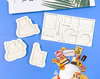 Puede incluir: Un juego de moldes de silicona blanca para hacer decoraciones de pasteles. Los moldes presentan varios vehículos de construcción, señales de tráfico y herramientas. Los moldes se muestran junto a un pastel decorado con figuras y señales de fondant con temática de construcción.