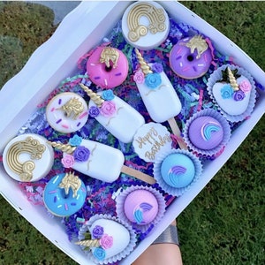 Pode incluir: Uma caixa branca cheia de guloseimas com tema de unic&oacute;rnio. A caixa cont&eacute;m donuts, cake pops e picol&eacute;s, todos decorados com chifres dourados, flores e detalhes de arco-&iacute;ris. O texto "Happy Birthday" tamb&eacute;m &eacute; vis&iacute;vel.