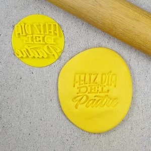 Puede incluir: Arcilla polimérica amarilla con el texto "Feliz día del Padre" impreso en ella. La arcilla está sobre una superficie gris junto a un rodillo de madera.