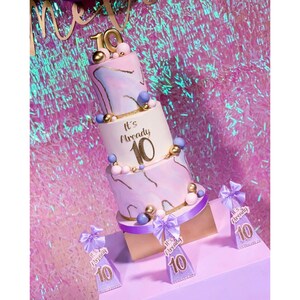 Pode incluir: Bolo de anivers&aacute;rio de tr&ecirc;s camadas com cobertura com efeito m&aacute;rmore rosa e roxo, detalhes dourados e o texto "It's Already 10". O bolo &eacute; decorado com esferas douradas e azuis. Inclui toppers decorativos a condizer.