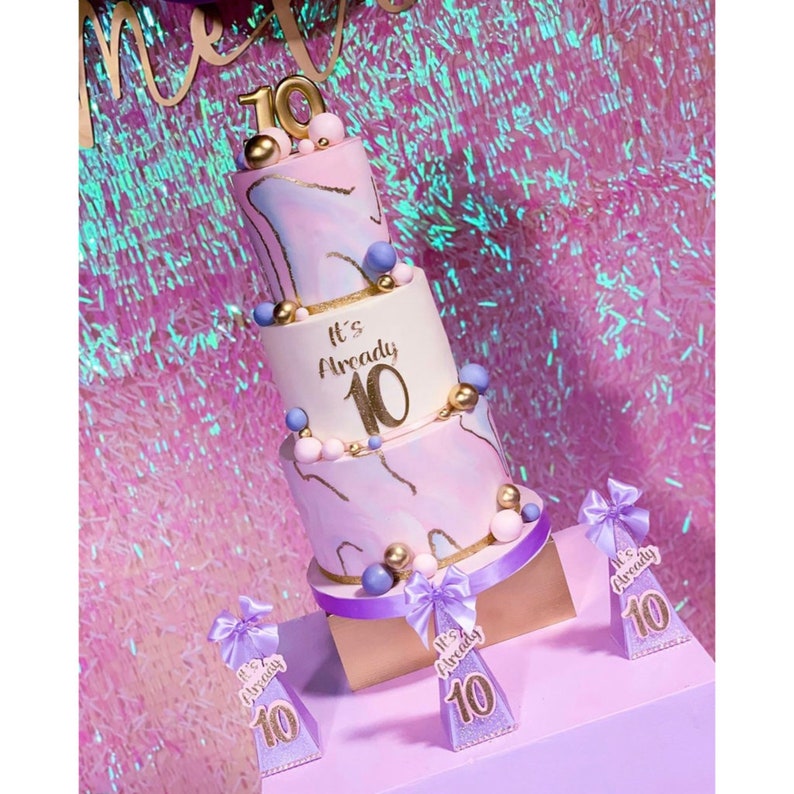 Pode incluir: Bolo de anivers&aacute;rio de tr&ecirc;s camadas com cobertura com efeito m&aacute;rmore rosa e roxo, detalhes dourados e o texto "It's Already 10". O bolo &eacute; decorado com esferas douradas e azuis. Inclui toppers decorativos a condizer.