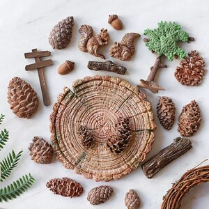 May include: A collection of miniature woodland-themed decorations. Includes brown pine cones, acorns, squirrels, a tree, a wooden sign, and a tree stump. The items are arranged on a white surface, creating a rustic and natural aesthetic.