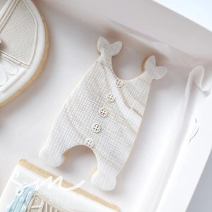 May include: A white cookie decorated to look like a baby onesie with a checkered pattern and white buttons. The cookie is on a white background.