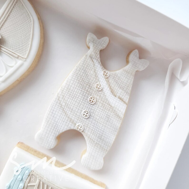 May include: A white cookie decorated to look like a baby onesie with a checkered pattern and white buttons. The cookie is on a white background.