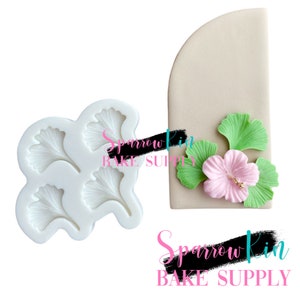 May include: A silicone mold for making fondant or gumpaste leaves. The mold features four leaf shapes, each with a detailed vein pattern. The mold is white and is shown next to a sample of the leaves made with the mold.