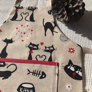 Peut inclure: Un tablier beige avec un motif de chat noir. Le tablier a une poche rouge avec une arête de poisson et un bol qui dit "kitty".