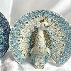 May include: Two ceramic peacock figurines with blue and green feathers and gold accents. The peacock on the left is blue and the peacock on the right is white.