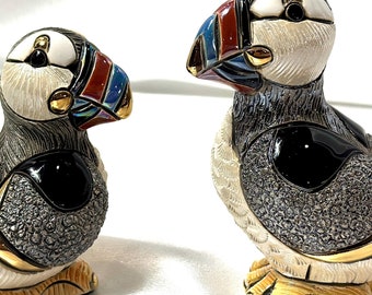 May include: Two ceramic figurines of puffins, one larger than the other. Both are painted in shades of gray, black, white, red, blue, and gold. The larger puffin is facing right, while the smaller puffin is facing left. Both have black eyes and gold accents.