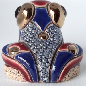 May include: A blue and red ceramic frog figurine with gold accents. The frog has a textured back and is sitting on a base.
