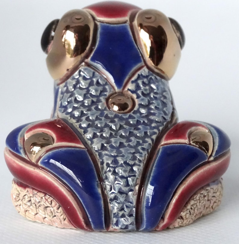 May include: A blue and red ceramic frog figurine with gold accents. The frog has a textured back and is sitting on a base.