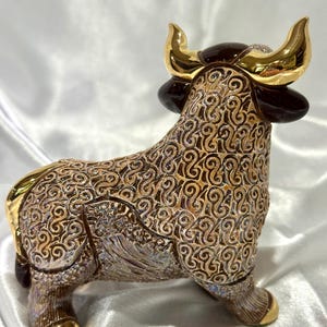 May include: A ceramic bull figurine featuring a detailed brown and tan swirling pattern across its body. The bull has polished gold-colored horns and hooves, with dark brown accents on the ears and head, standing in a profile orientation.